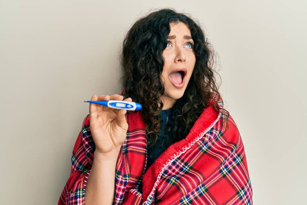 Young brunette woman with curly hair wrapped in a blanket holding thermometer angry and mad screaming frustrated and furious, shouting with anger looking up.  - Foto, Imagen