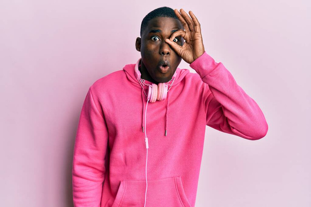 Mladý africký Američan, oblečený do tělocvičny a používající sluchátka, dělá v pořádku gesto šokované překvapeným obličejem, oči hledící skrz prsty. nevěřící výraz.  - Fotografie, Obrázek