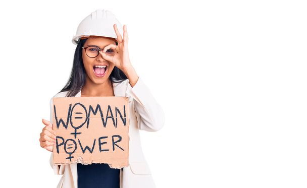 Young beautiful latin girl wearing architect hardhat holding woman power banner smiling happy doing ok sign with hand on eye looking through fingers  - Foto, afbeelding