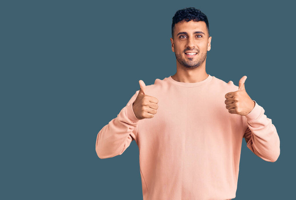 Jonge Spaanse man draagt casual kleding succes teken doet positief gebaar met de hand, duimen omhoog glimlachend en gelukkig. vrolijke uitdrukking en winnaar gebaar.  - Foto, afbeelding