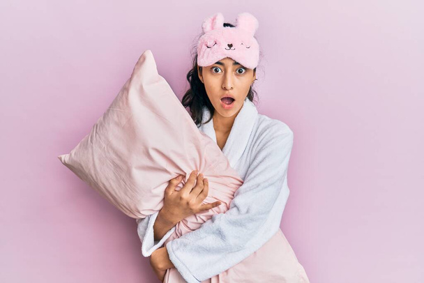Hispanic teenager girl with dental braces wearing sleep mask and robe hugging pillow afraid and shocked with surprise and amazed expression, fear and excited face.  - Photo, Image