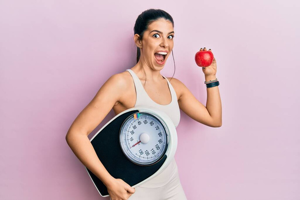Νεαρή ισπανίδα γυναίκα φορώντας αθλητικά κρατώντας μηχανή ζύγισης και μήλο γιορτάζει τρελό και έκπληκτος για την επιτυχία με ανοιχτά μάτια ουρλιάζοντας ενθουσιασμένος.  - Φωτογραφία, εικόνα