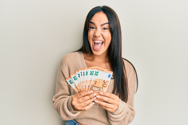 Young beautiful hispanic girl holding bunch of 50 euro banknotes celebrating crazy and amazed for success with open eyes screaming excited.  - Fotó, kép