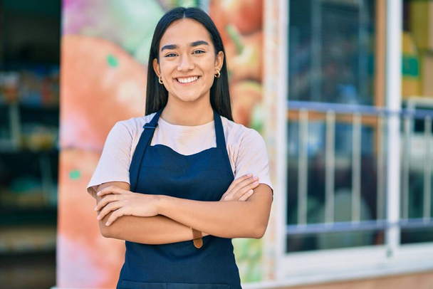 Young latin shopkeeper girl with arms crossed smiling happy at the fruit store. - 写真・画像