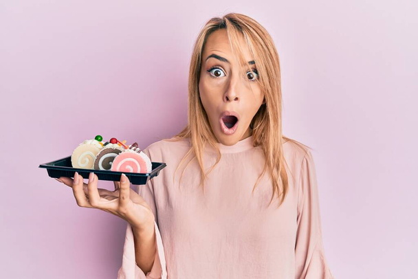 Krásná blondýnka drží dort sladkosti vyděšené a ohromený s otevřenými ústy pro překvapení, nevěřící tvář  - Fotografie, Obrázek