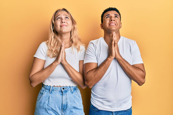 Молодая межрасовая пара в обычной белой футболке просит и молится руками вместе с выражением надежды на лице очень эмоциональным и беспокойным. begging.  - Фото, изображение