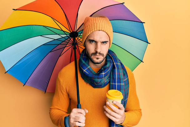 Homem hispânico jovem segurando colorido guarda-chuva beber tirar café cético e nervoso, franzindo a testa chateado por causa do problema. pessoa negativa.  - Foto, Imagem
