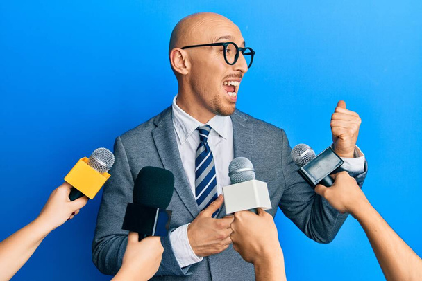 Bald man with beard being interviewed by reporters holding microphones pointing thumb up to the side smiling happy with open mouth  - 写真・画像