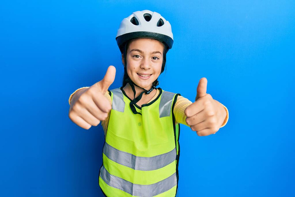 Красивая брюнетка маленькая девочка в велосипедном шлеме и рефлексивный жилет одобряющий делать позитивный жест с рукой, большие пальцы вверх улыбается и счастлив за успех. жест победителя.  - Фото, изображение