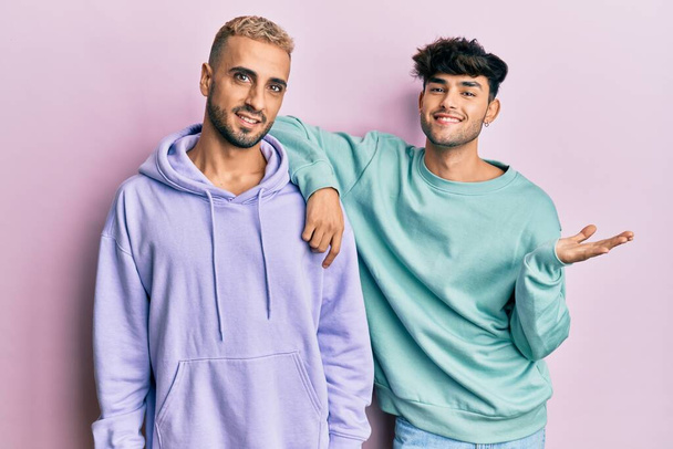 Homosexual gay couple standing together wearing casual clothes smiling cheerful presenting and pointing with palm of hand looking at the camera.  - Photo, Image