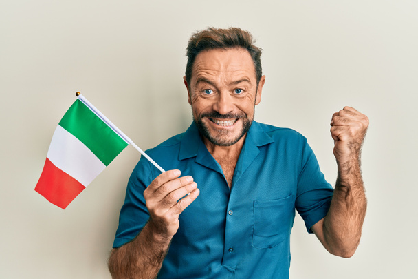 Mężczyzna w średnim wieku trzyma włoską flagę krzycząc dumny, świętując zwycięstwo i sukces bardzo podekscytowany podniesionym ramieniem  - Zdjęcie, obraz