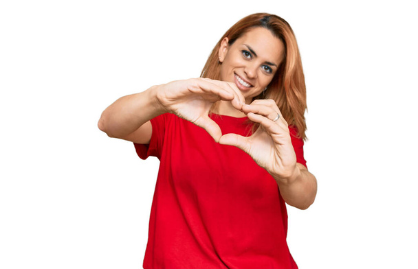 Jeune femme hispanique portant un t-shirt rouge décontracté souriant en amour faisant forme de symbole de coeur avec les mains. concept romantique.  - Photo, image