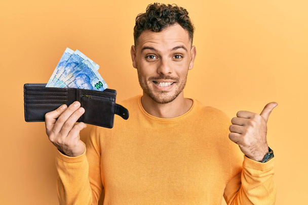 Молодий іспаномовний чоловік тримає гаманець з південноафриканськими банкнотами рандо, вказуючи великий палець в сторону посміхаючись з відкритим ротом
  - Фото, зображення
