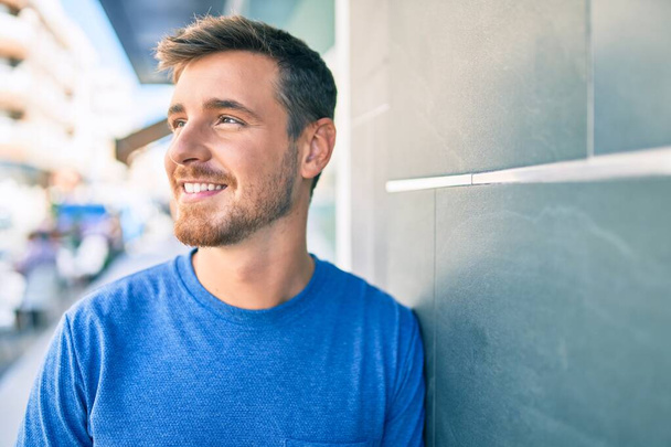 Fiatal fehér férfi mosolyog boldogan támaszkodik a falra a városban. - Fotó, kép
