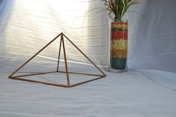 Piramida miedziana z oświetleniem, z zabytkowym szklanym wazonem i kamieniami w widoku bocznym, z naturalnym oświetleniem tła, piramida do użytku domowego - Zdjęcie, obraz
