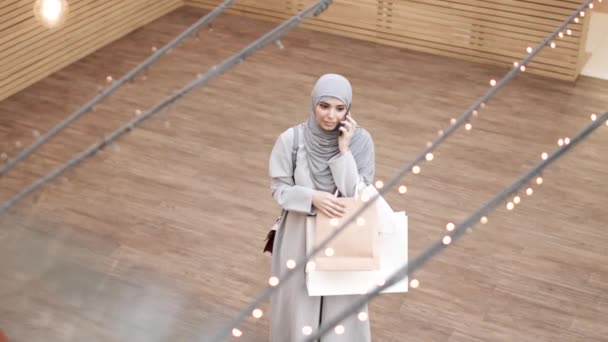 Vista de alto ângulo da jovem mulher muçulmana encantadora vestindo abaya cinza e hijab segurando sacos de compras em pé dentro de casa, conversando no telefone celular, em seguida, olhando para a câmera e acenando com a mão - Filmagem, Vídeo