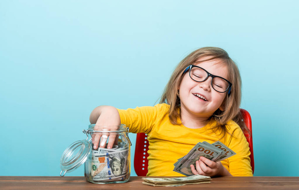 Hymyilevä pikkutyttö ottaa rahaa lasipurkista, se on täynnä euroja. Sen vieressä on kasa kolikoita ja säästöpossu. Valtava määrä rahaa on kertynyt. Lahjoittaa hyväntekeväisyyteen - Valokuva, kuva