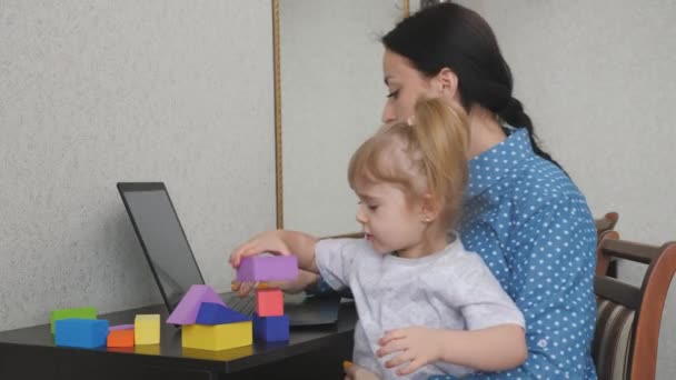Mamma lavora in un computer portatile con un bambino piccolo tra le braccia, il bambino raccoglie una torre da cubi colorati, una donna freelance si prende cura del bambino, lavora da casa, seduto a casa, fa soldi online - Filmati, video