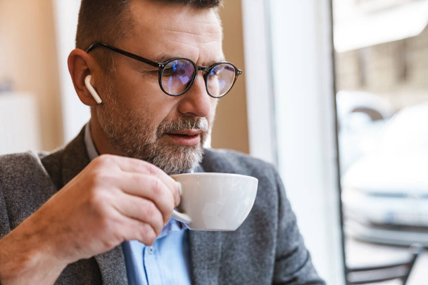 Close up of a handsome confident businessman having breakfast in modern coffee shop, drinking coffee, wearing wireless earphones - Fotoğraf, Görsel