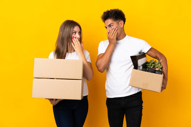 Teenager coppia che si trasferisce in una nuova casa tra scatole isolate su sfondo blu che copre la bocca con le mani per dire qualcosa di inappropriato - Foto, immagini