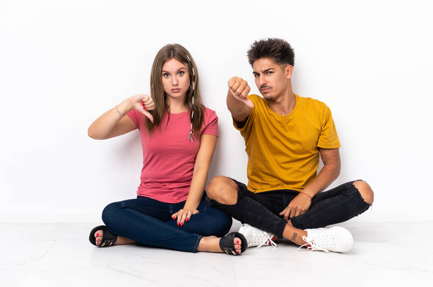 Young couple sitting on the floor isolated on white background showing thumb down with negative expression - Fotó, kép