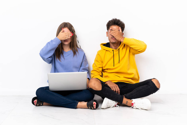 白い背景に手で目をカバーに隔離された床に座ってノートパソコンを持つ若いカップル。何か見たくない - 写真・画像