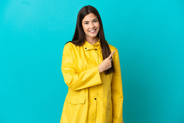 Подростковая бразильская девушка в непромокаемом пальто на изолированном синем фоне, указывая на сторону, чтобы представить продукт - Фото, изображение