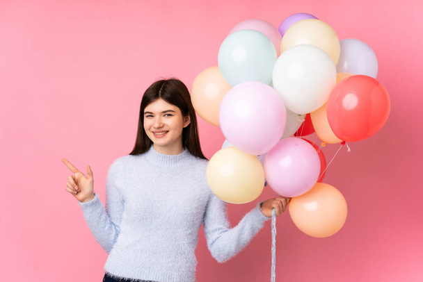 Young Ukrainian teenager girl holding lots of balloons over isolated pink background pointing finger to the side - Photo, Image