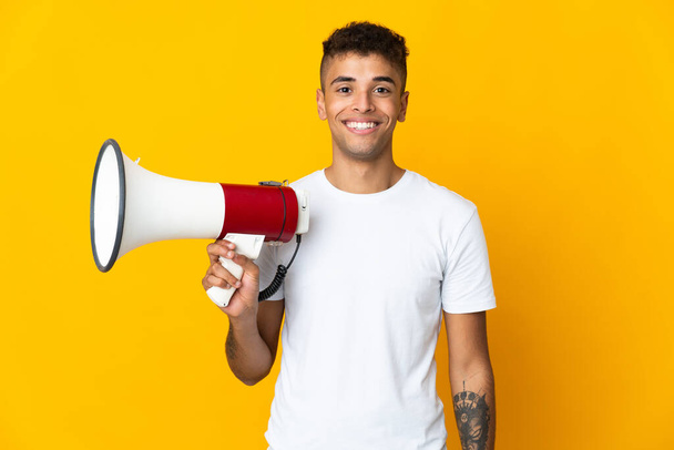 Young brazilian man isolated on yellow background holding a megaphone and smiling a lot - Photo, Image