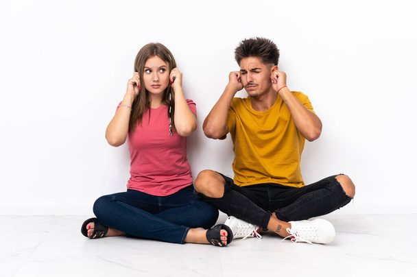 Young couple sitting on the floor isolated on white background frustrated and covering ears - Foto, Imagem