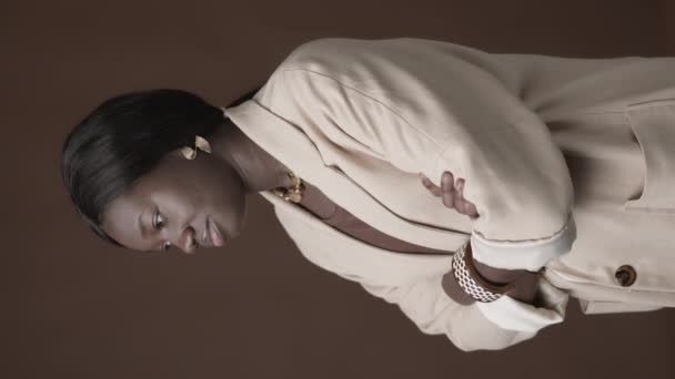 Vue verticale de la jeune femme afro-américaine portant des vêtements et accessoires élégants debout sur fond brun avec les bras croisés sur la poitrine et regardant la caméra - Séquence, vidéo