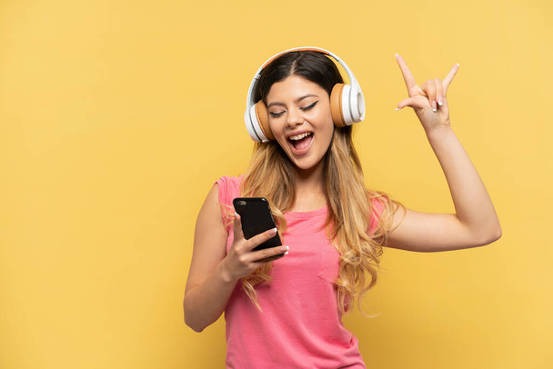 Молодая россиянка, изолированная на желтом фоне, слушает музыку с помощью мобильного рок-жеста - Фото, изображение