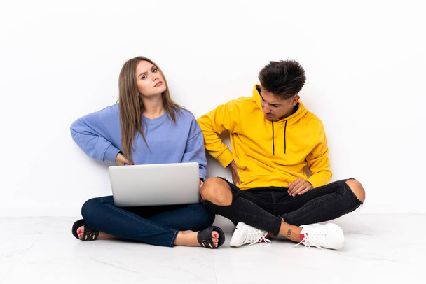 Giovane coppia con un computer portatile seduto sul pavimento isolato su sfondo bianco che soffre di mal di schiena per aver fatto uno sforzo - Foto, immagini