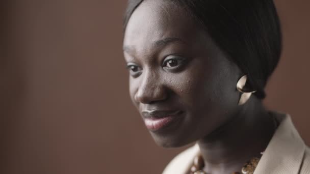 Gros plan portrait de jeune femme afro-américaine portant des vêtements décontractés et de beaux accessoires debout sur fond brun et regardant de côté - Séquence, vidéo