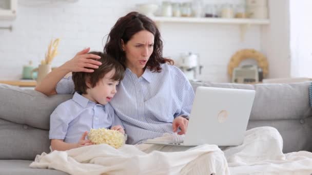Mom and kid son with bowl of popcorn watching scary movie closing their eyes sitting on sofa at home - Footage, Video