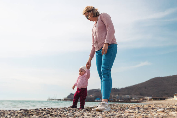 Matka trzyma dziecko za rękę i spaceruje z nim po plaży, wzdłuż morza. Outdoor Walking na świeżym powietrzu. - Zdjęcie, obraz