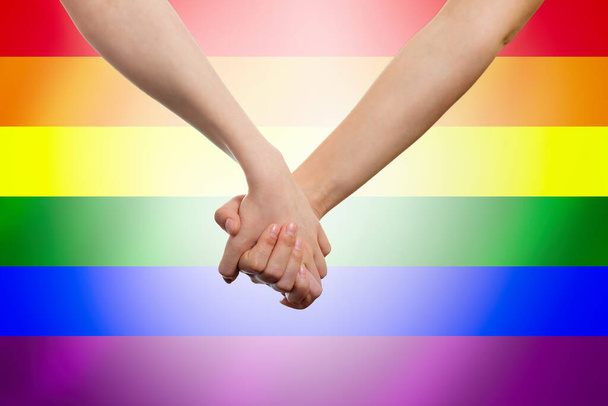 Female's hands hold each other against the background of a rainbow LGBT flag. Close-up. The concept of LGBT equality. - Photo, Image