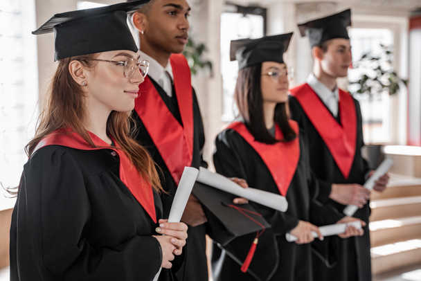 estudiante feliz cerca de compañeros de clase interracial en vestidos de graduación y gorras con diploma sobre fondo borroso  - Foto, Imagen