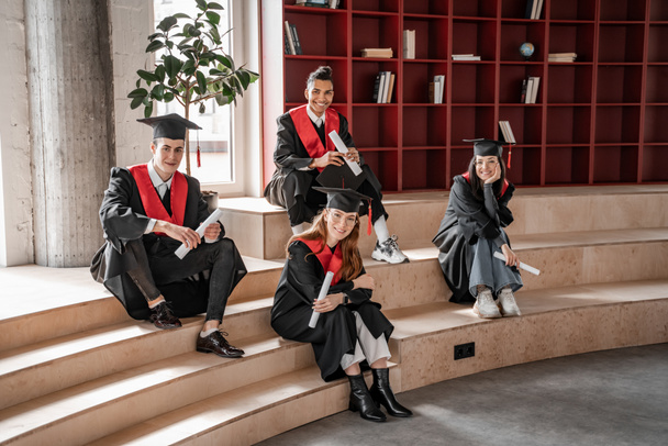 estudiantes interracial feliz en batas y gorras con diploma y sentado en las escaleras, la clase de graduación 2021 - Foto, Imagen
