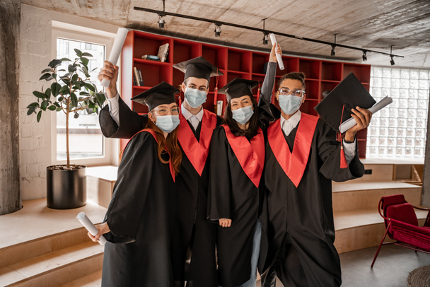estudiantes multiétnicos en máscaras médicas, vestidos y gorras con diploma, clase de graduación 2021 - Foto, Imagen