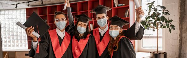 estudiantes multiétnicos en máscaras médicas, vestidos de graduación y gorras con diploma, pancarta - Foto, Imagen