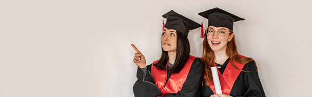 bastante estudiante en vestido de graduación y gorra señalando con el dedo cerca compañero de clase alegre, senior 2021, pancarta - Foto, Imagen