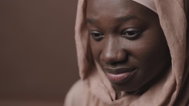 Acercamiento de la cara de una joven musulmana africana con un hermoso pañuelo en la cabeza de pie sobre un fondo marrón y mirando a un lado - Metraje, vídeo