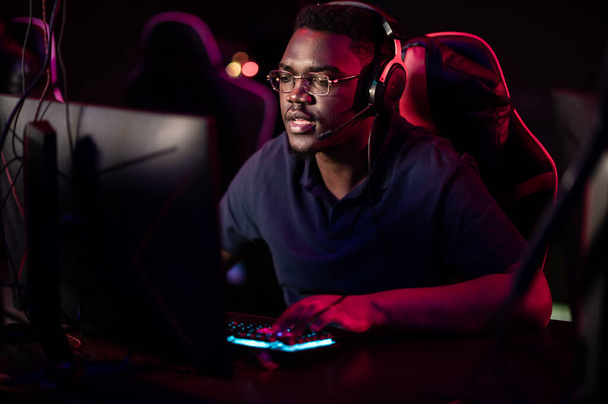 Un estudiante africano se sienta en una silla de juego en un club de computadoras, se pone auriculares, sonríe y se prepara para el juego - Foto, imagen