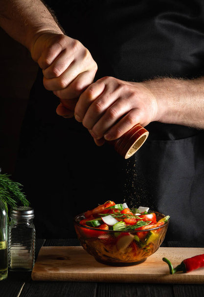 Restoran mutfağındaki şef taze sebze salatasına biber ekler. Elinde değirmen tutan bir aşçının yakın çekimi. Sağlıklı ve lezzetli yemekler pişirmek. - Fotoğraf, Görsel