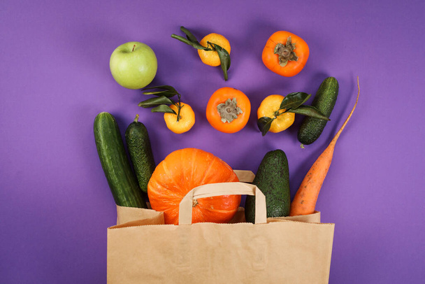 Green and orange fruits and vegetables in the craft paper bag on the violet background. Top view. Grocery shopping. Persimmon, tangerines, cucumbers, apple, carrot and pumpkin. Vegetarian food.  - Fotografie, Obrázek