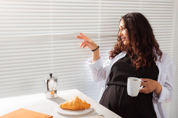 Радостная беременная молодая красивая женщина смотрит сквозь жалюзи во время утреннего завтрака с кофе и круассанами. Концепция доброго утра и ожидания встречи с ребенком. Copyspace - Фото, изображение
