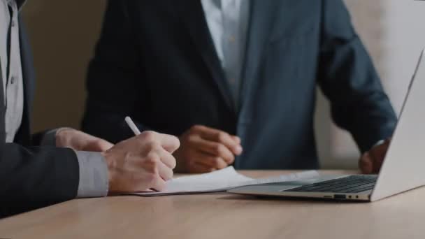 Close-up two multiethnic unrecognizable business men sitting in office with papers sign contract agreement shake hands. Male hands signing report agreement making partnership union gesture handshake - Footage, Video