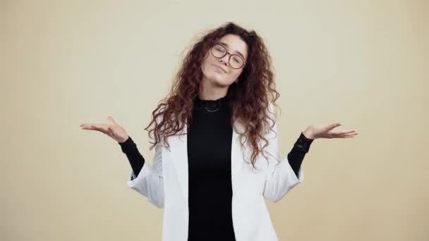 The smiling young woman with curly hair, holds her hands to the side and says look like that, smiling wide with her teeth. Young hipster in gray jacket and white shirt, with glasses posing isolated on - Footage, Video