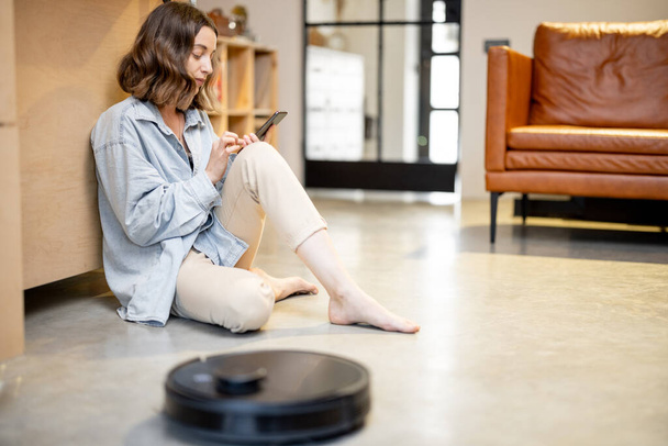 Relaxed woman with phone and robotic vacuum cleaner - Fotoğraf, Görsel